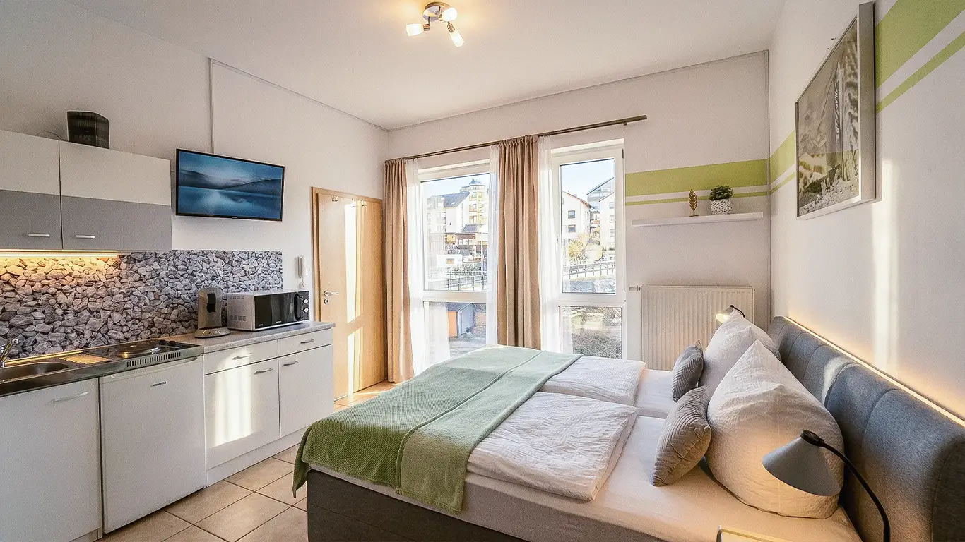 Modernes Gästezimmer mit Doppelbett, grüner Decke, Küchenzeile und großen Fenstern mit Blick auf umliegende Gebäude.