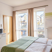 Helles Schlafzimmer mit Doppelbett, grüner Decke und Blick auf Häuser durch ein großes Fenster.