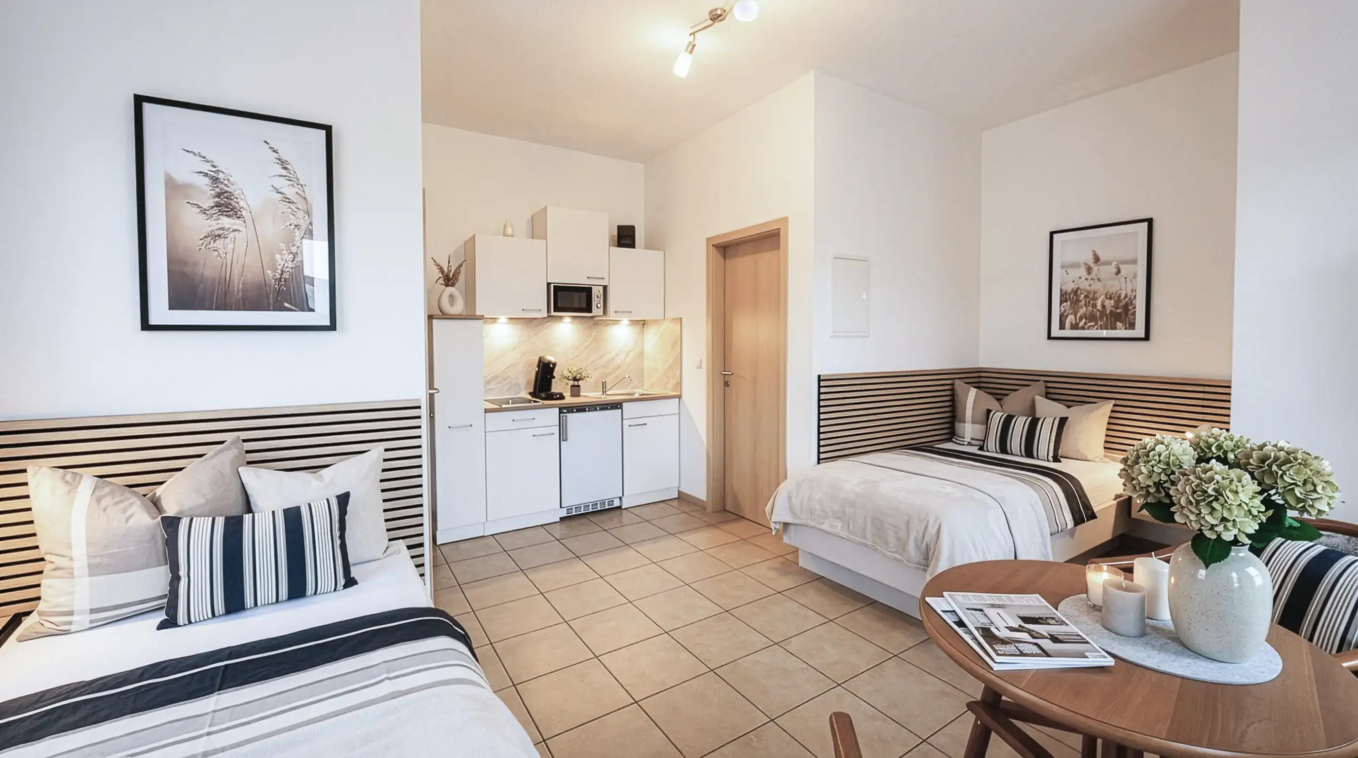 Helles, modernes Zimmer mit zwei Einzelbetten, Küchenzeile und einem kleinen Holztisch mit Vase, Kerzen und Zeitschriften.
