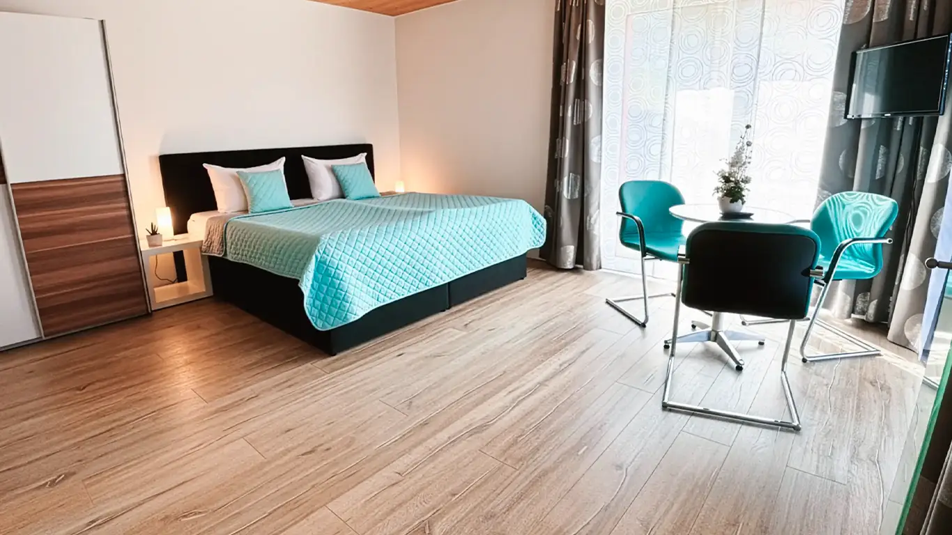 Modernes Schlafzimmer mit großem Bett in türkisfarbenem Bettüberwurf, bequemen Sesseln um einen runden Glastisch und einem Wandfernseher.