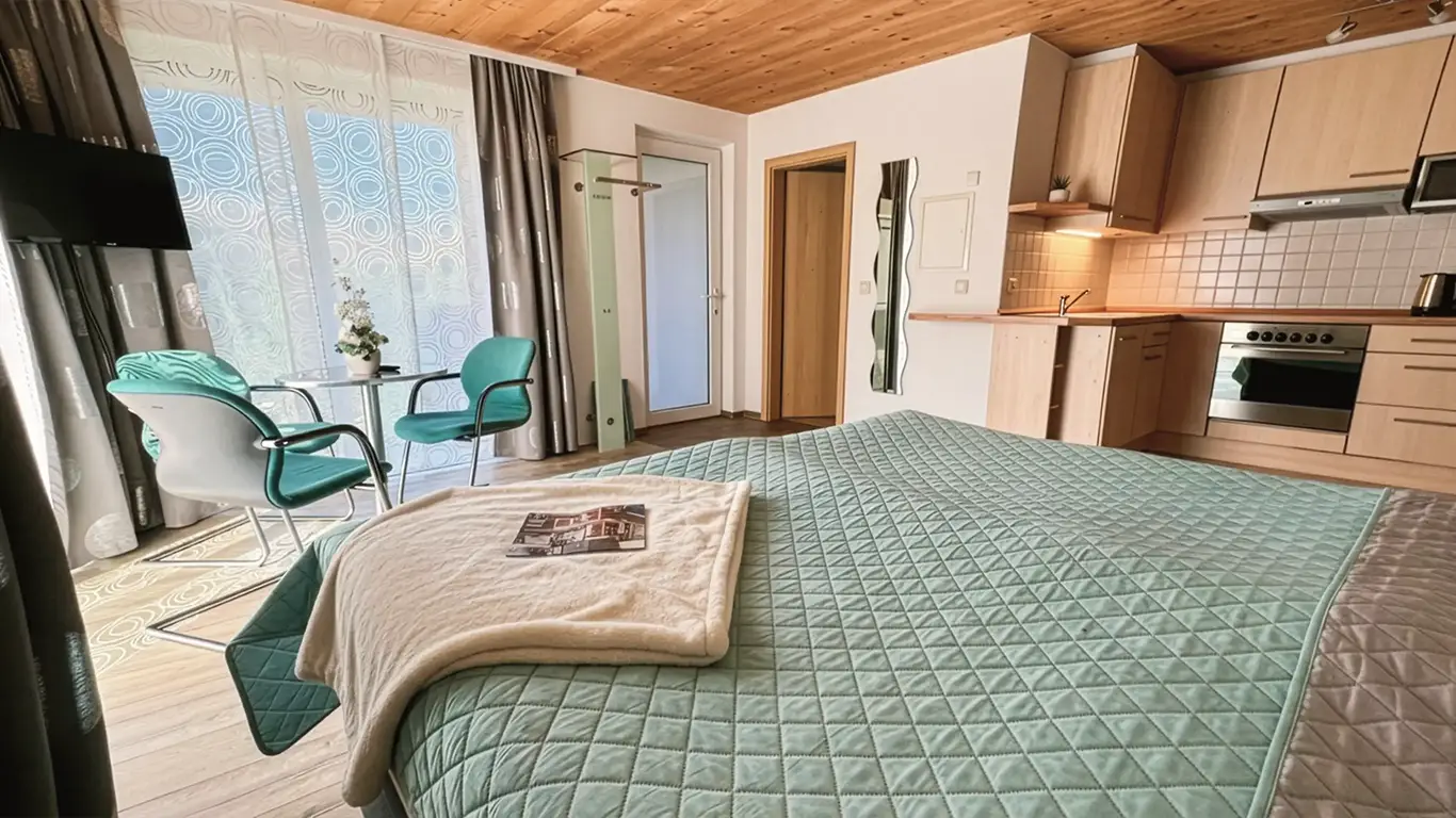 Modernes Schlafzimmer mit grün gesteppter Bettdecke, zwei grünen Stühlen am Fenster und offener Küchenzeile.