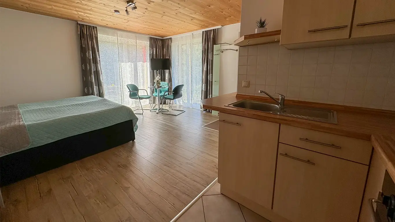 Helles Apartment mit Holzboden, Bett mit blauem Überwurf, kleiner Sitzecke vor bodentiefen Fenstern und Küchenzeile mit Spüle.