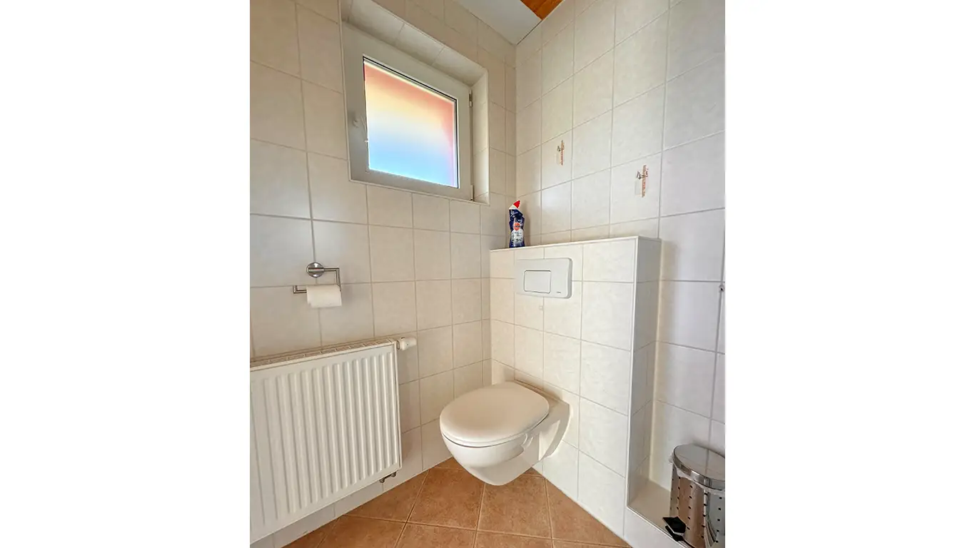 Moderner, weiß gefliester Badezimmerbereich mit wandhängendem WC, Heizkörper, kleiner Fenster und Abfalleimer.