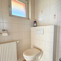 Kleines, weiß gekacheltes Badezimmer mit WC, Heizkörper, Fenster mit farbiger Folie und Toilettenpapierhalter.