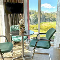 Gemütlicher Sitzbereich mit zwei grünen Stühlen an einem kleinen runden Tisch vor einem großen Fenster mit Blick auf eine grüne Wiese und Bäume.