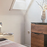 Helles Schlafzimmer mit Schrägenfenster, Nachttischlampe, Holzkommode und Vase mit Trockenblumen.