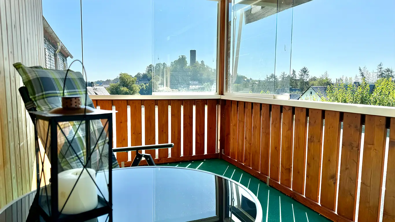 Balkon mit Holzgeländer, grün kariertem Stuhl, Laterne mit Kerze und Blick auf einen Turm bei klarem Himmel.