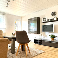 Helles Wohnzimmer mit Holzboden, einem modernen Esstisch, drei Stühlen, TV an der Wand und Dekorationen auf Regalen.