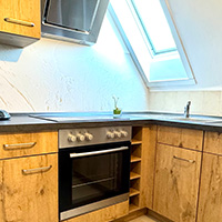 Helle Küchenzeile mit Holzschränken, Elektroherd, Spüle unter Dachfenster und kleinem Pflanzen-Topf auf der Arbeitsplatte.