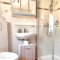 Kleines Badezimmer mit weißem Waschbecken, Holzunterschrank, Spiegel und Dusche mit Glasabtrennung.