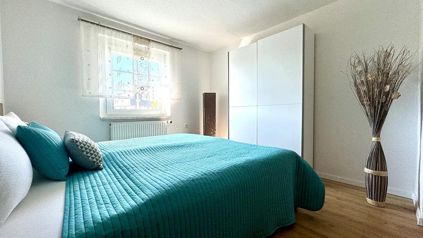 Helles Schlafzimmer mit Bett in Türkis, zwei Kissen, weißem Kleiderschrank und großer Vase mit Trockenblumen.