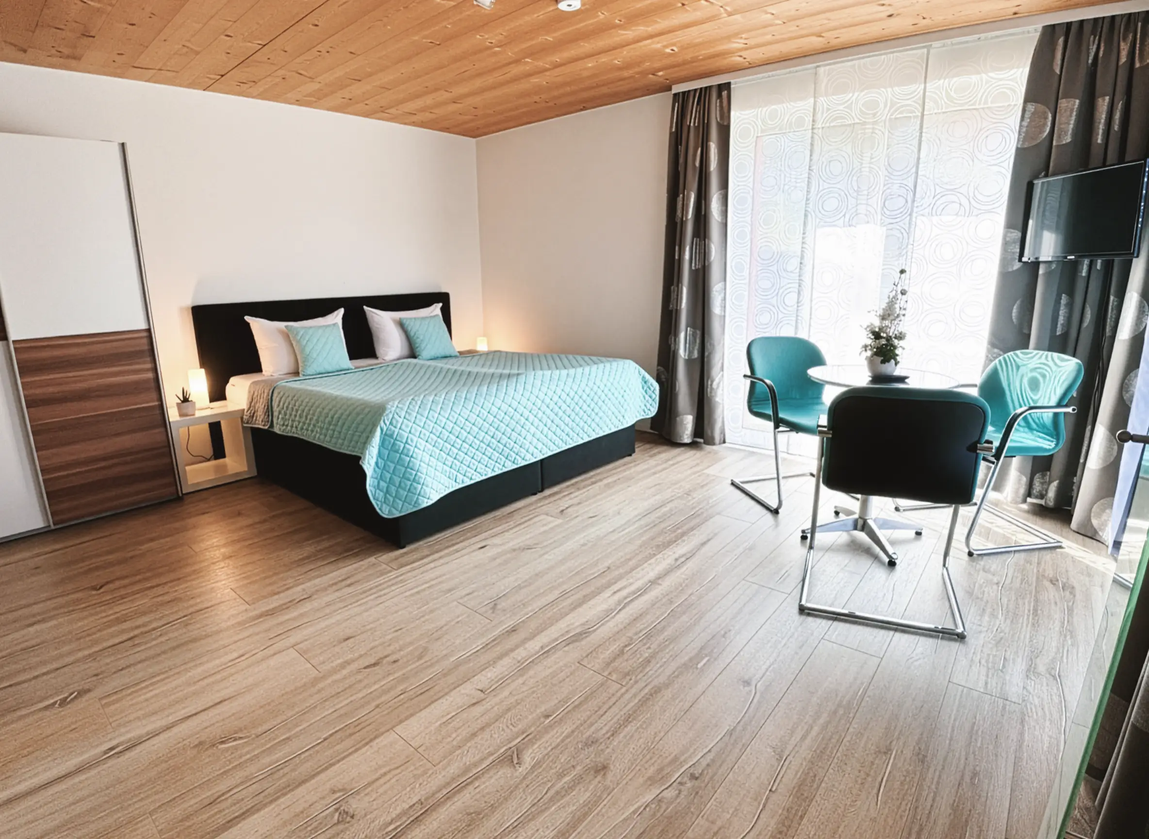 Helles Schlafzimmer mit Holzboden, Doppelbett mit türkiser Bettdecke und einem kleinen Tisch mit drei Stühlen vor einem großen Fenster.