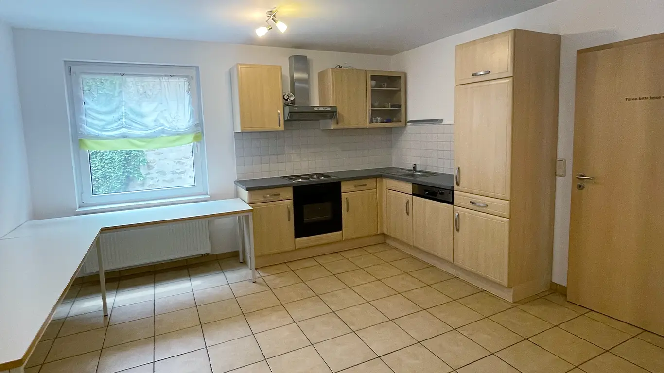 Kleine Küche mit hellen Holzschränken, schwarzer Arbeitsplatte, eingebautem Herd und Spüle sowie einem großen weißen Ecktisch unter einem Fenster mit weiß-grüner Gardine.