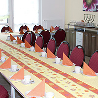 Ein lang gedeckter Tisch mit roten Stühlen, orangefarbenen gefalteten Servietten und Tee- oder Kaffeetassen in einem hellen Raum.