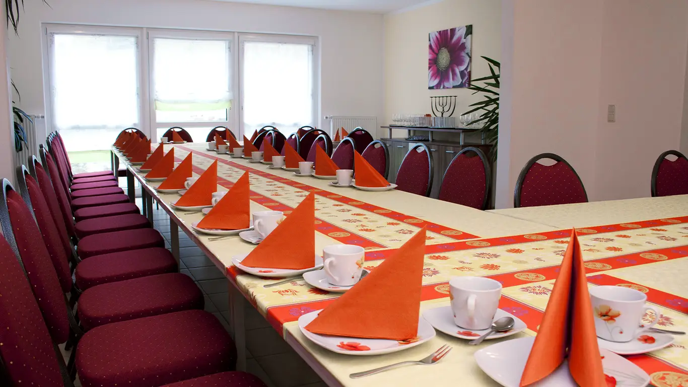 Langer Tisch mit rotgefalteten Servietten, Tassen und Tellern, umgeben von roten Stühlen in einem hellen Raum.
